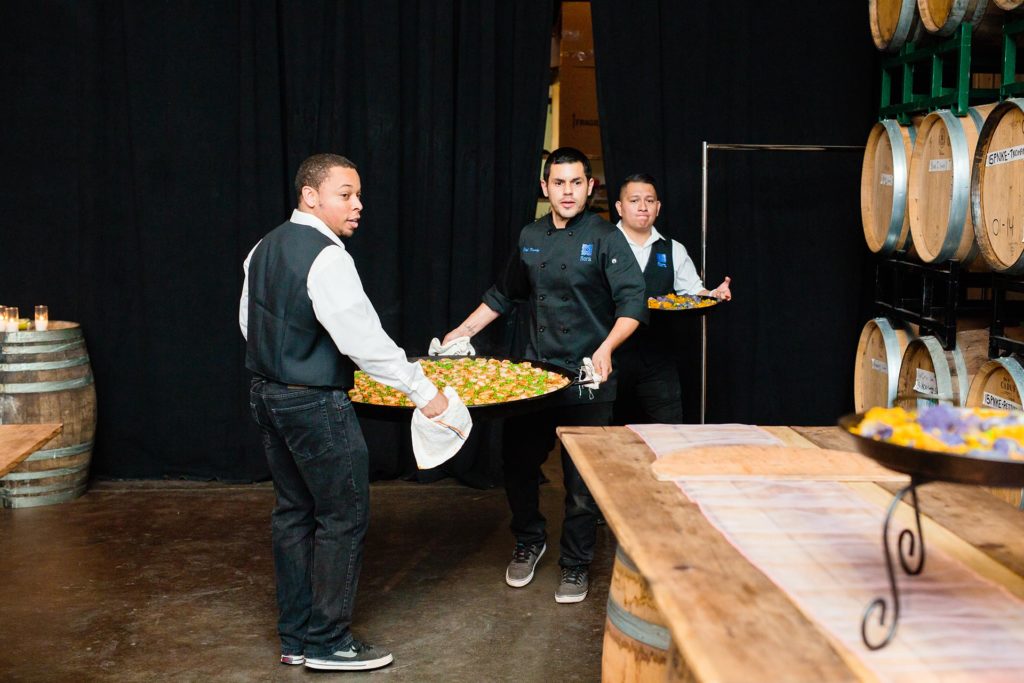 Catering staff carrying a large paella pan toward a buffet beside wine barrels.