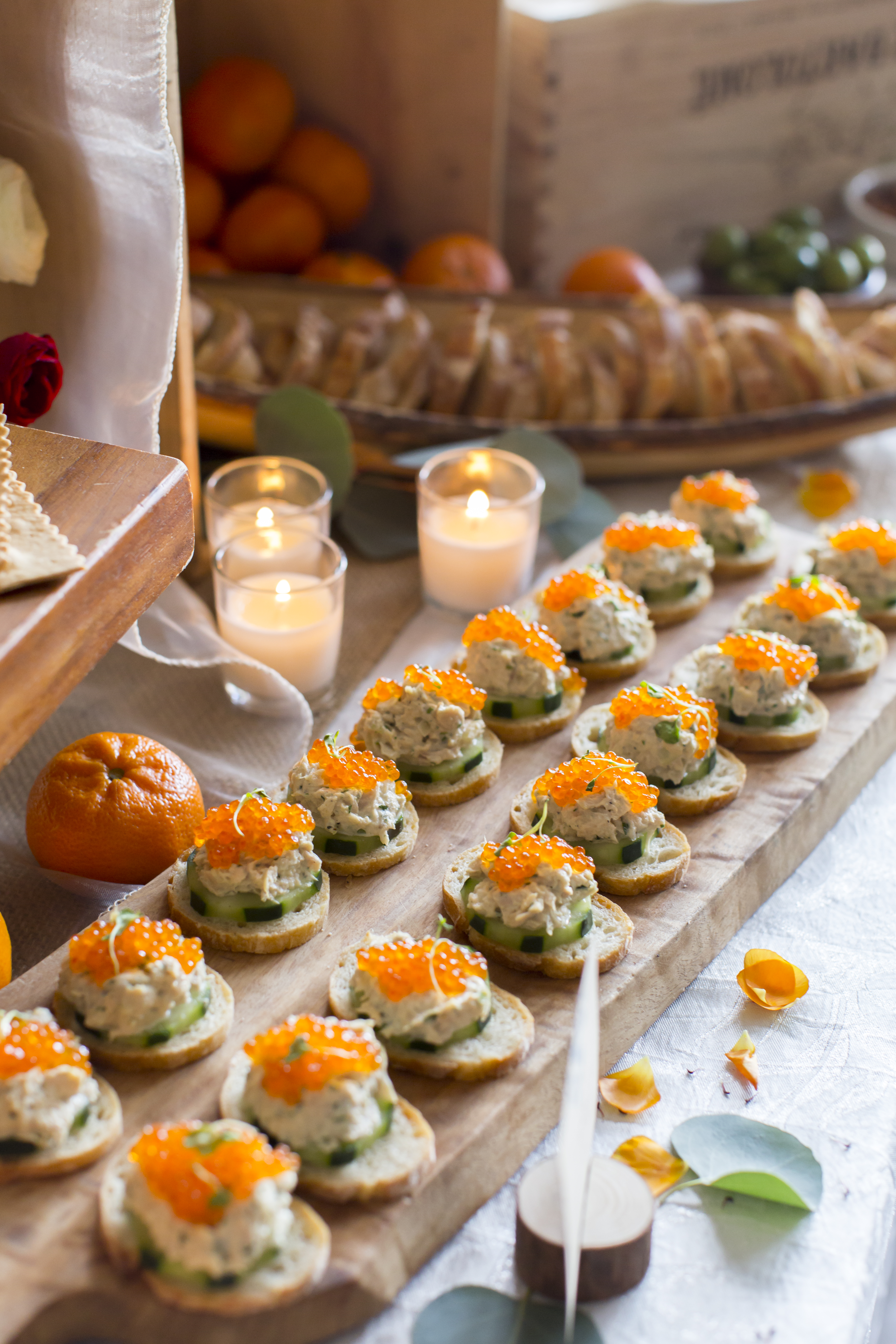 Crostini topped with cucumber, creamy salad and orange roe, arranged beside candles and citrus.