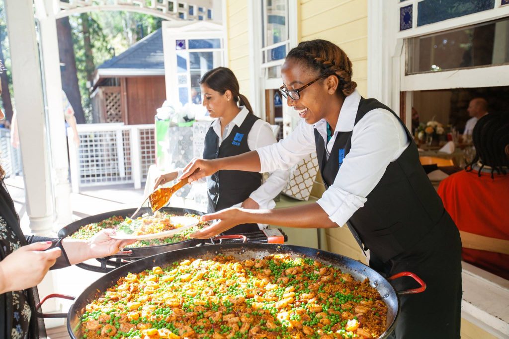 Two Ñora servers plating paella for guests at a reception buffet.