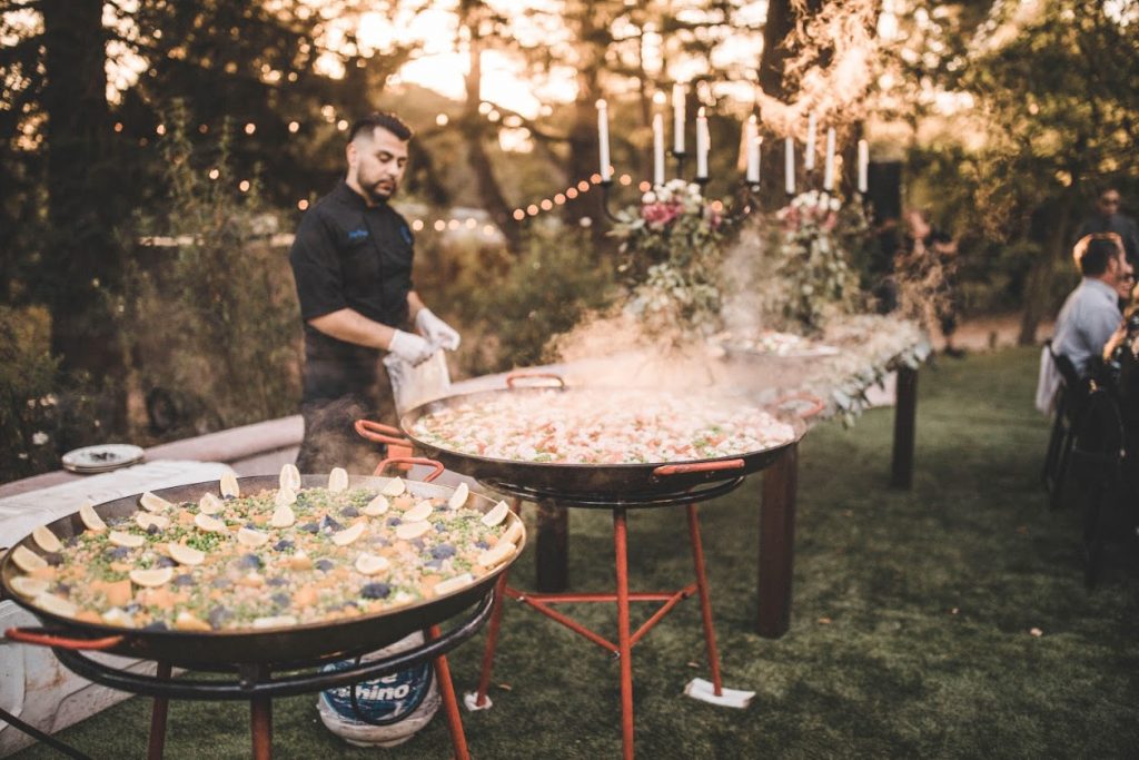 Ñora chef cooking paella outdoors beneath trees and string lights at sunset.