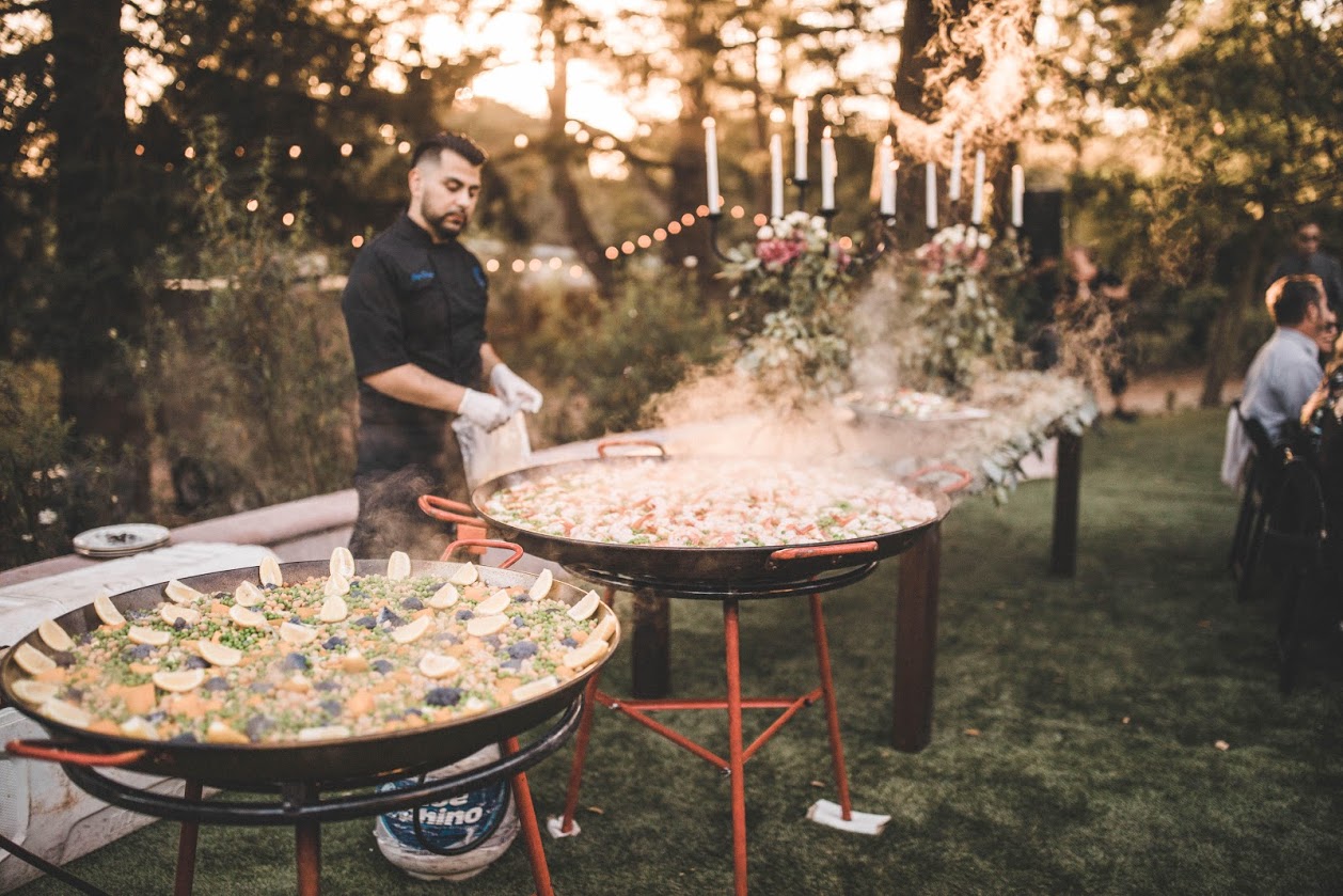 A chef tending paella pans beneath trees in the evening light.