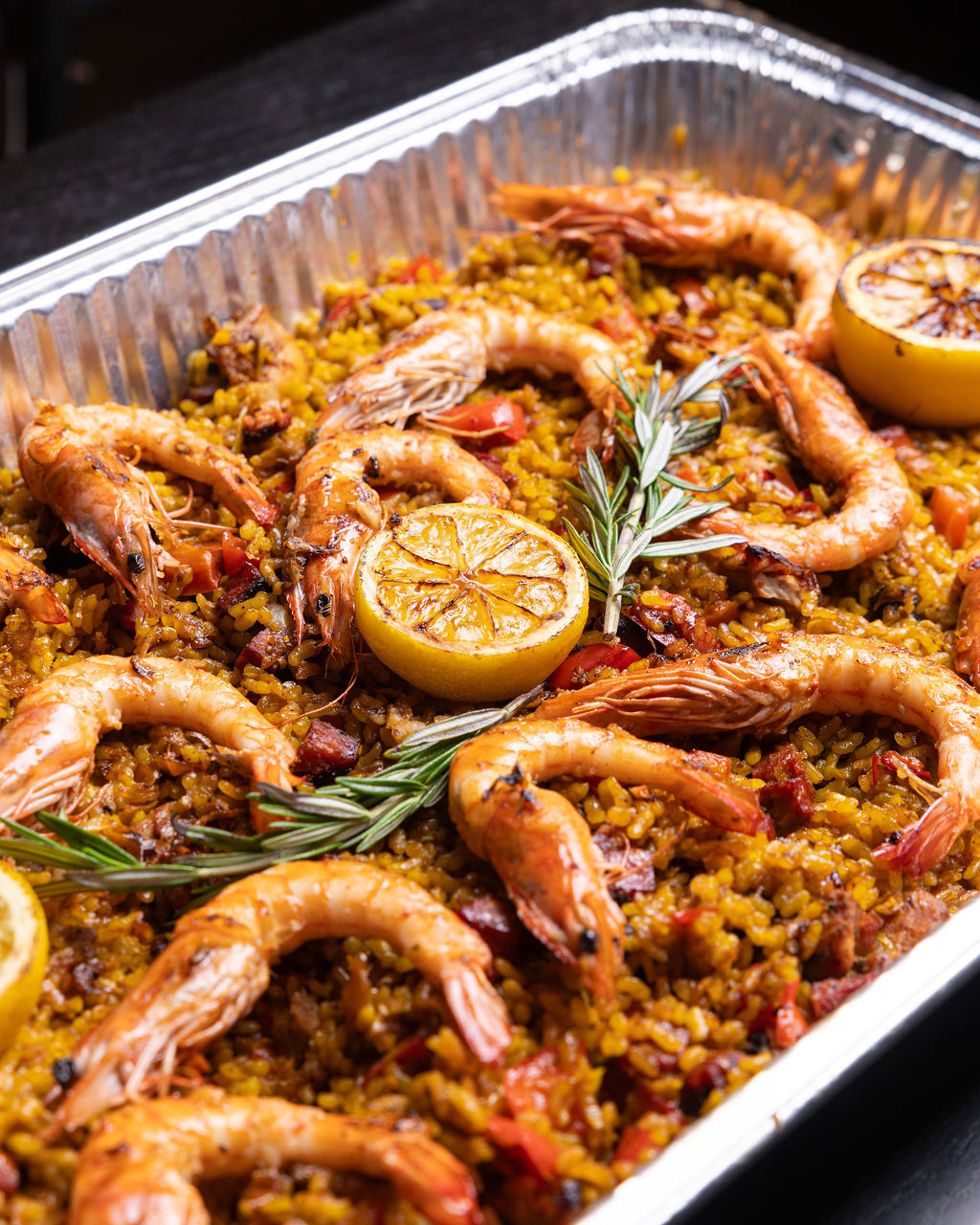 Shrimp paella in an aluminum catering tray with grilled lemon halves and rosemary.