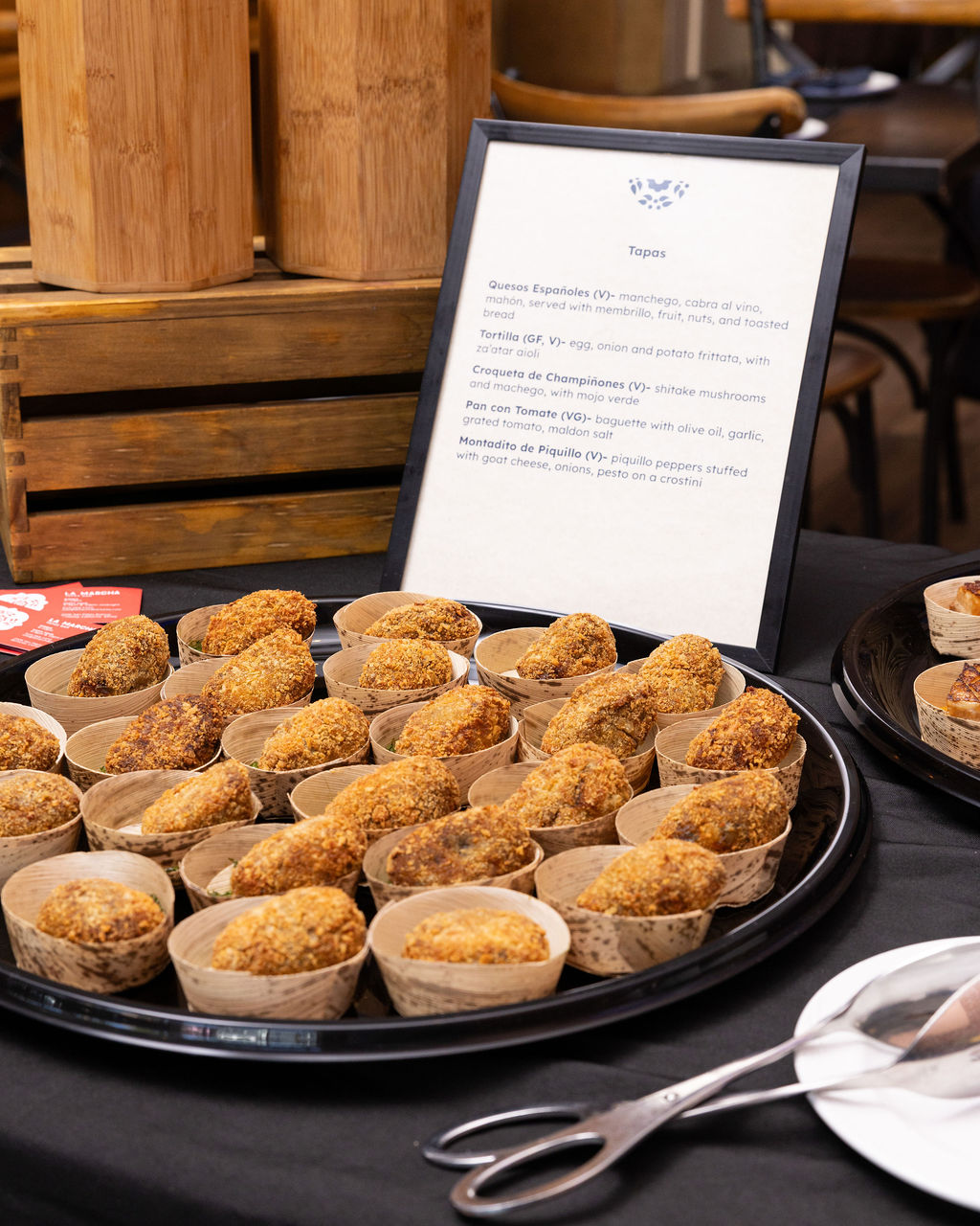 Golden croquetas in individual serving cups on a catering platter beside a tapas menu.